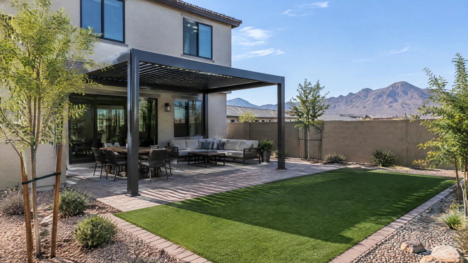 Backyard of a newer two-story stucco home in southwest Phoenix: a freestanding aluminum pergola over a paver patio, young trees, artificial turf, block wall, desert mountains far in the distance