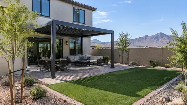 Backyard of a newer two-story stucco home in southwest Phoenix: a freestanding aluminum pergola over a paver patio, young trees, artificial turf, block wall, desert mountains far in the distance