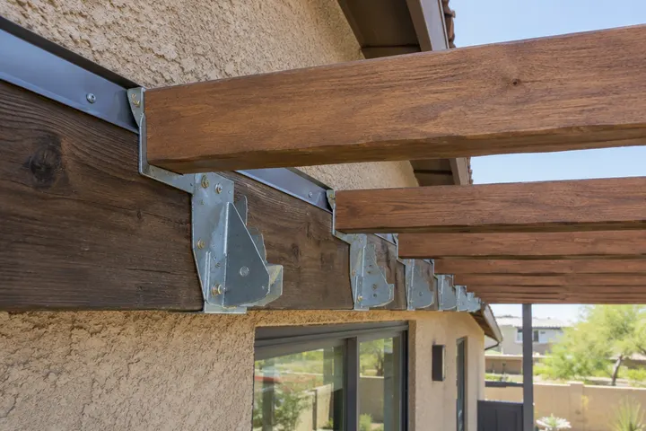 Metal rafter hangers on a ledger board at the house wall supporting pergola rafters, flashing above, close-up